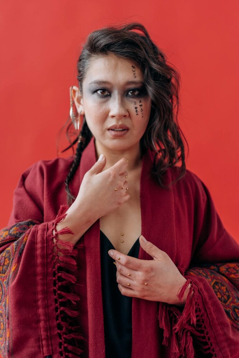 Woman in red shawl with artistic makeup poses confidently against a red backdrop.